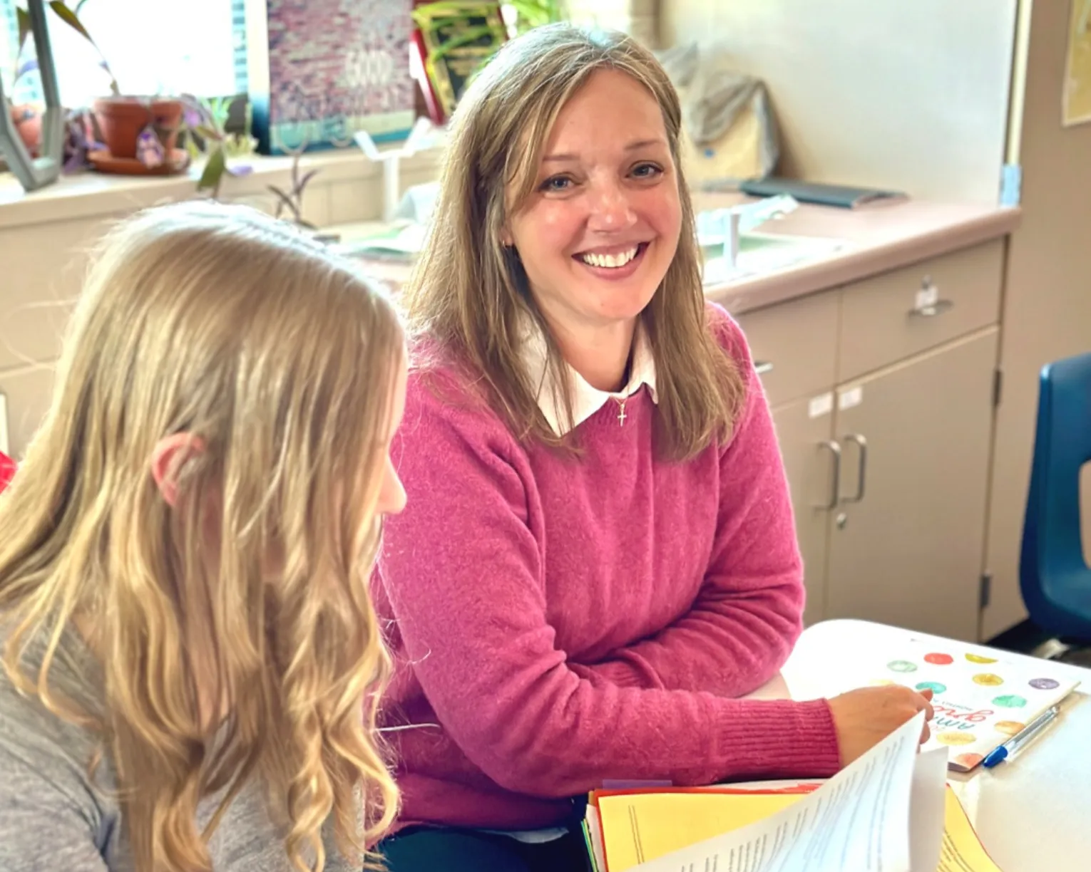 A member in a pink sweater looks up at the camera with a smile as she works with a student on a reading packet