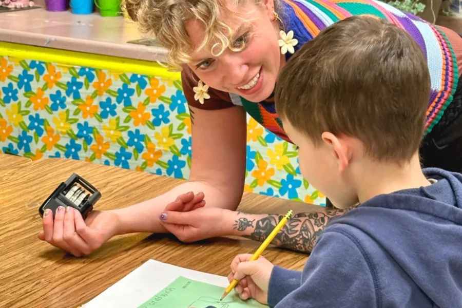 A WEAC member with flower earrings and curly blonde hair helps an elementary student at their desk